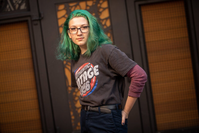 A girl with green hair stands with hands in back pocket.