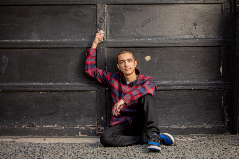 A boy in a plaid shirt sits against a black wall.