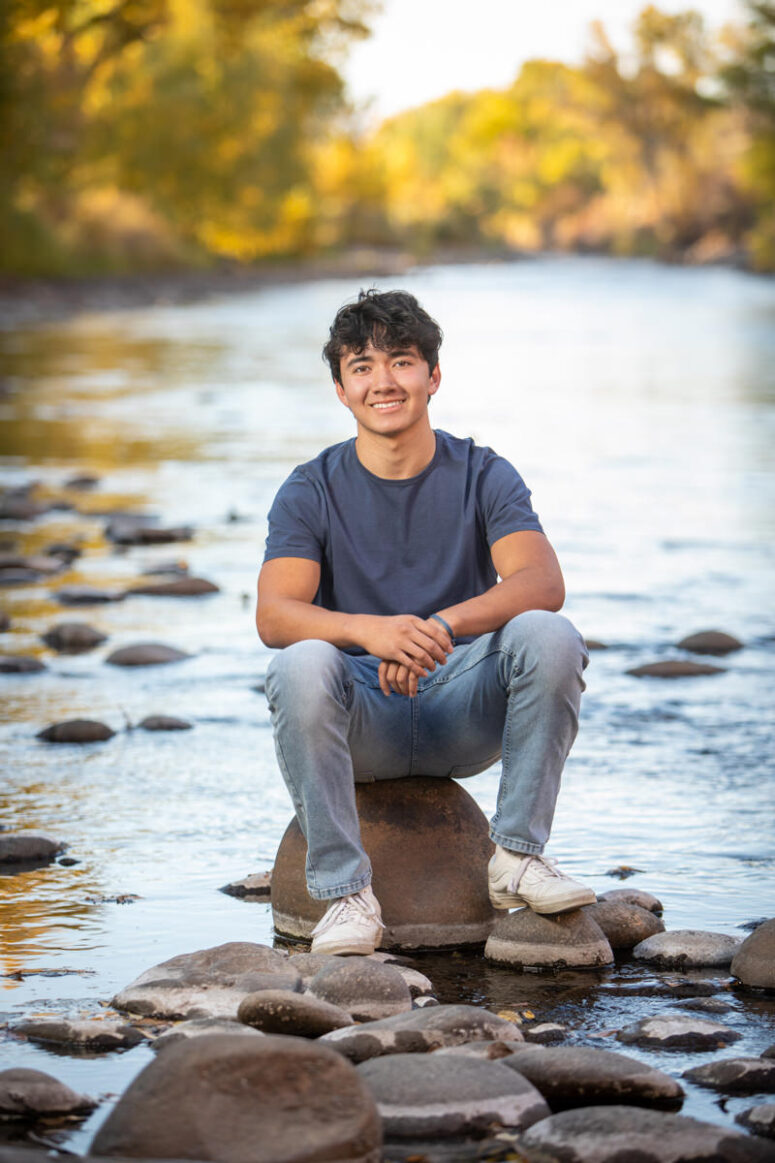 A guy in blue shirt sits on rock in river.