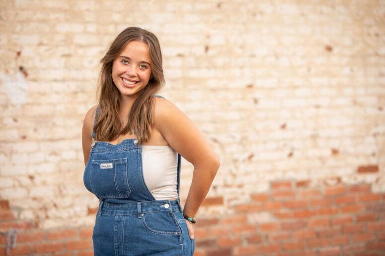 Senior portrait of girl in overalls against brick wall.