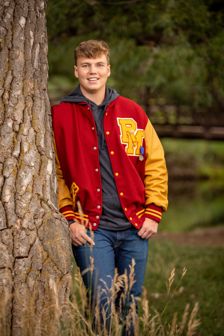 A football players stands against tree with lettermans jacket on.