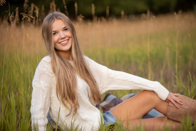 girl in white sweat smiling while sitting in tall grass.